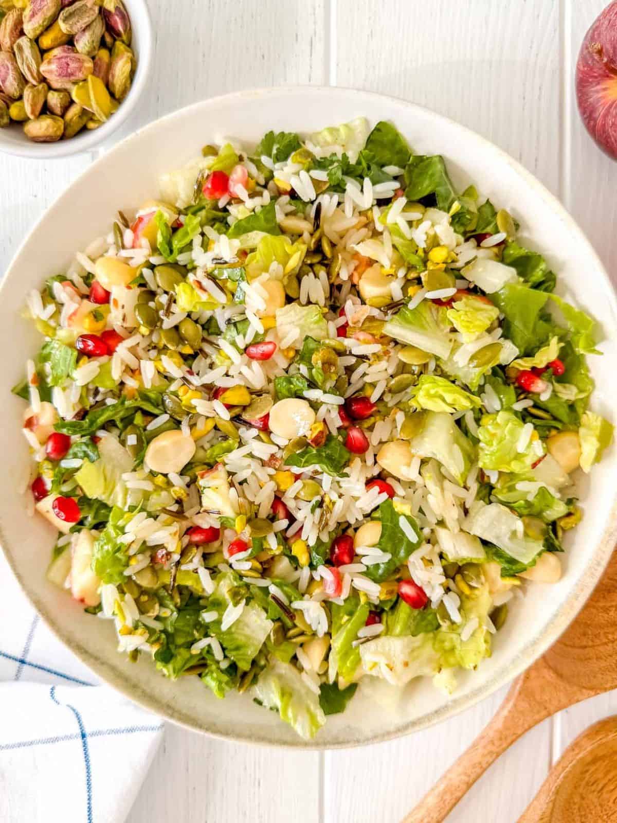 fruit and nut salad with rice in a white bowl next to salad servers, red apple and bowl of pistachios.