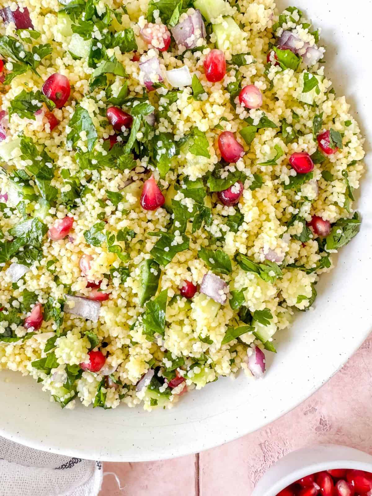 couscous tabbouleh in a white bowl next to a small bowl of pomegranate seeds.