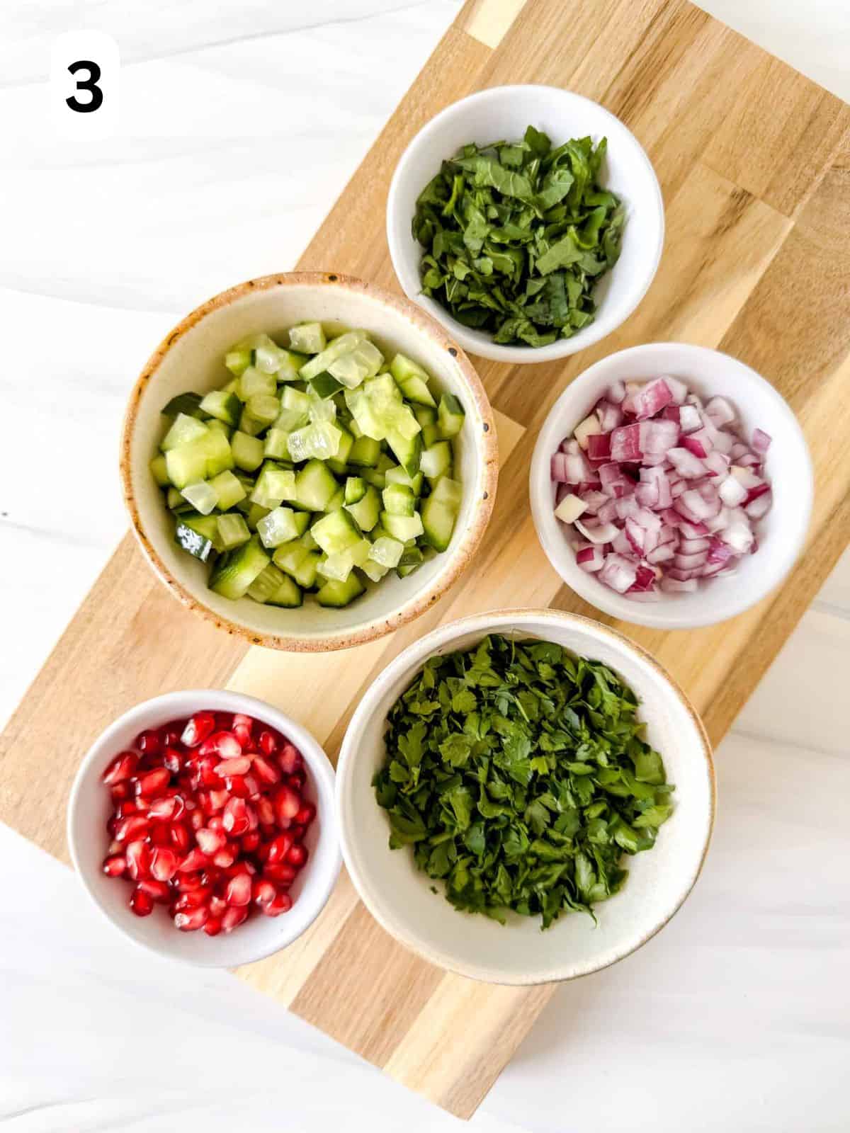 five small bowls of diced cucumber, red onion, fresh parsley, fresh mint leaves and pomegranate seeds labelled number three.