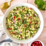 couscous tabbouleh in a white bowl on a pink background next to fresh parsley and pomegranate seeds.