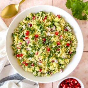 couscous tabbouleh in a white bowl on a pink background next to fresh parsley and pomegranate seeds.