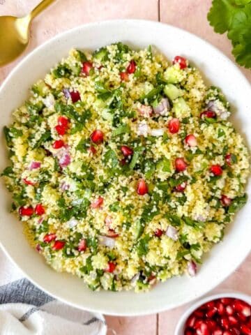 couscous tabbouleh in a white bowl on a pink background next to fresh parsley and pomegranate seeds.