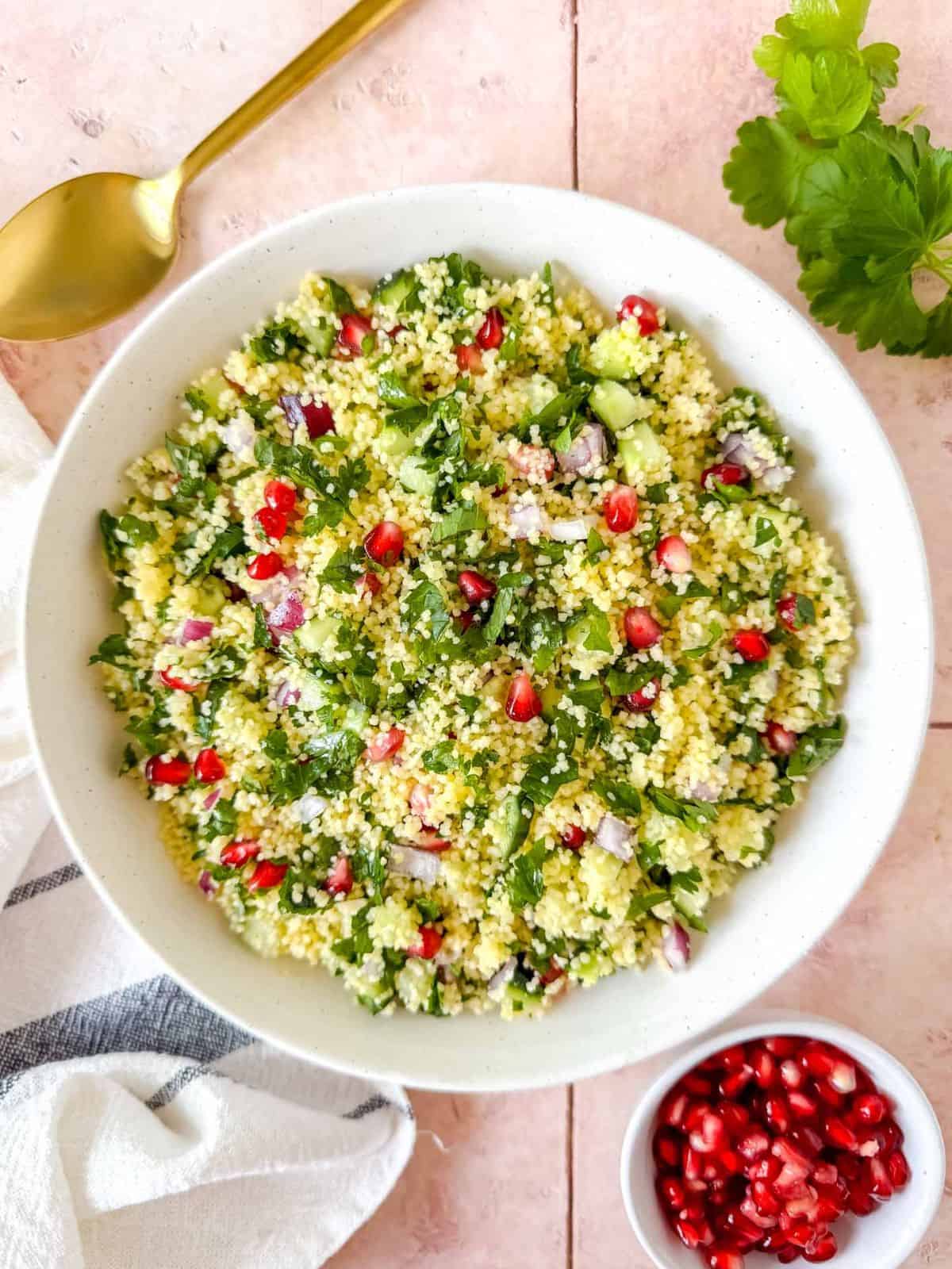 couscous tabbouleh in a white bowl on a pink background next to fresh parsley and pomegranate seeds.