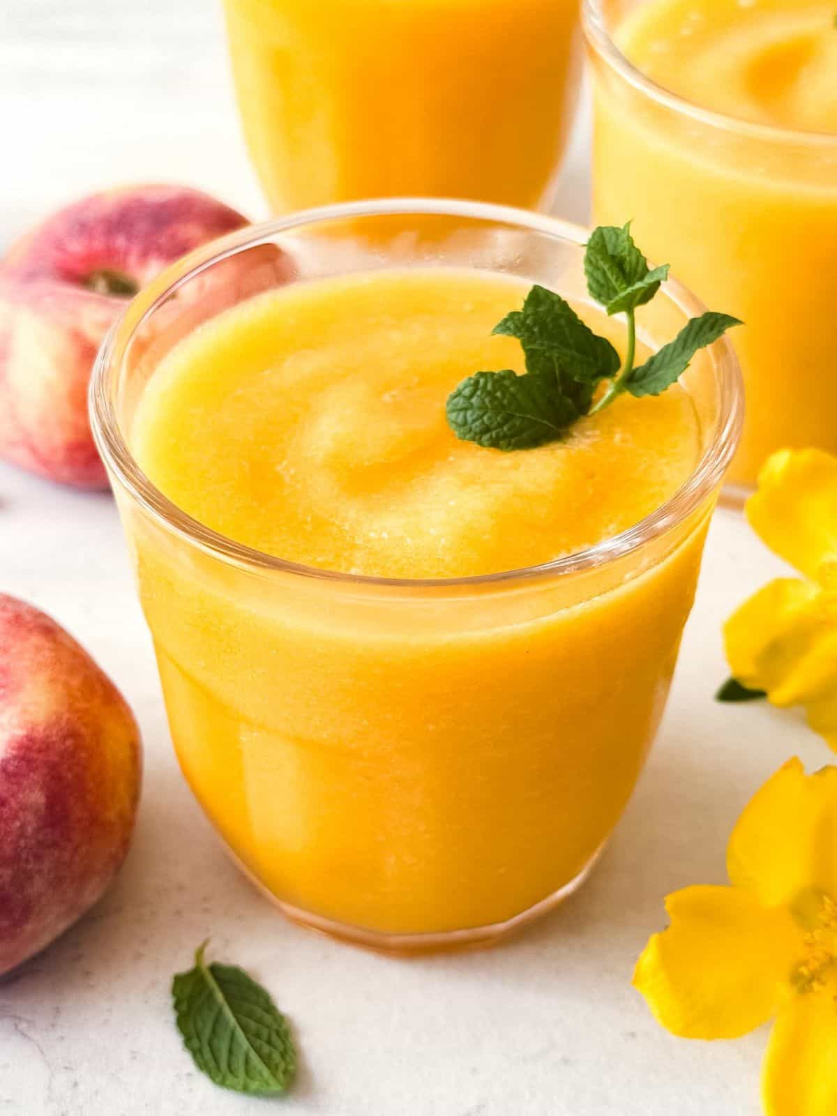 frozen peach slush without wine in three glasses next to fresh peaches and mint leaves.
