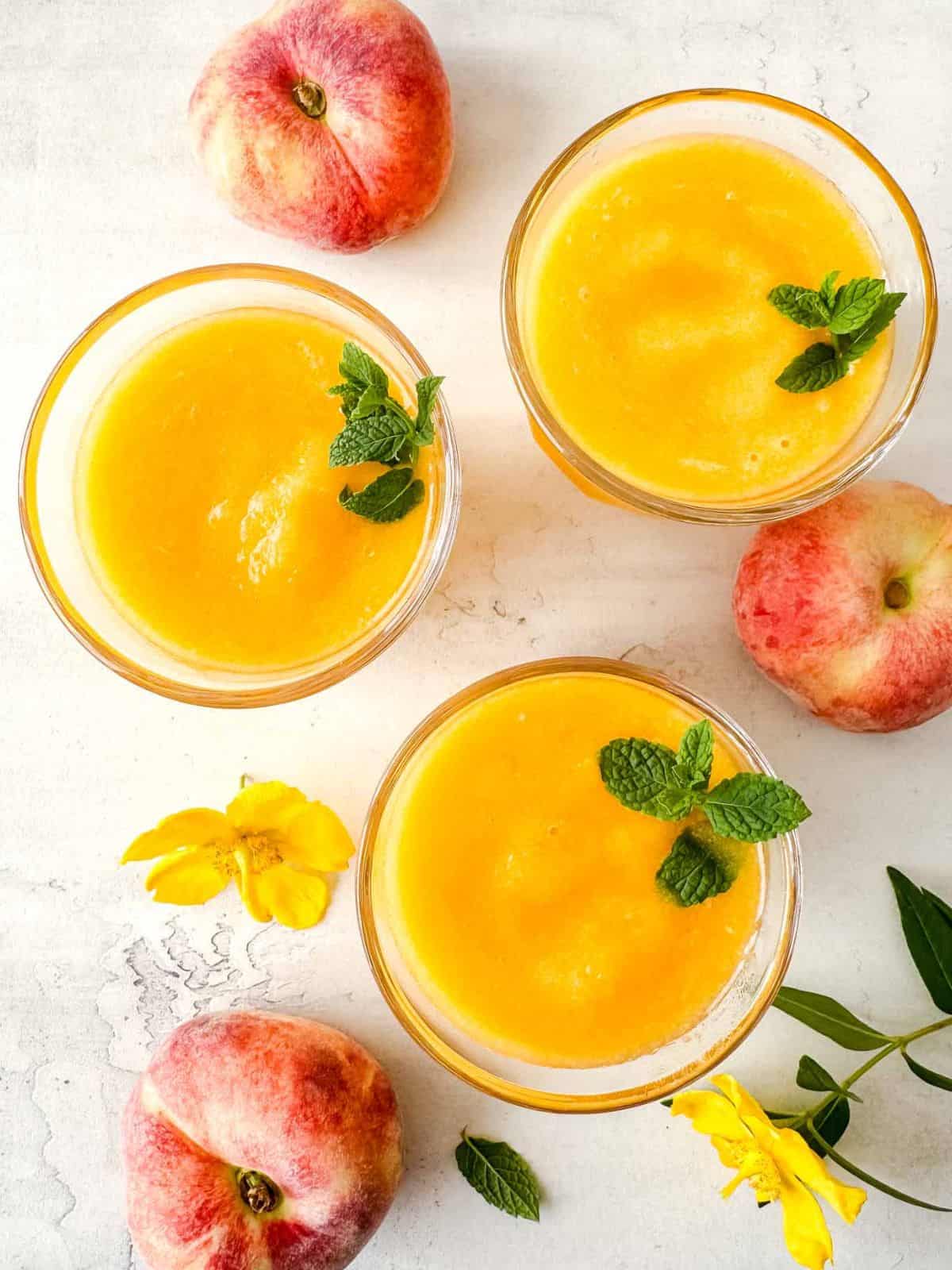 three glasses of frozen peach slush drink next to fresh peaches and yellow flowers.