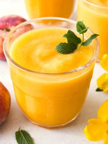 frozen peach slush without wine next to fresh peaches, mint leaves and yellow flowers.