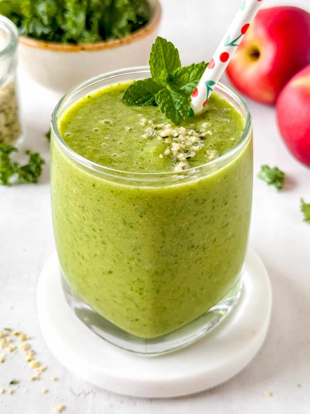 green low histamine smoothie in a glass next to fresh fruit, kale and hemp seeds.