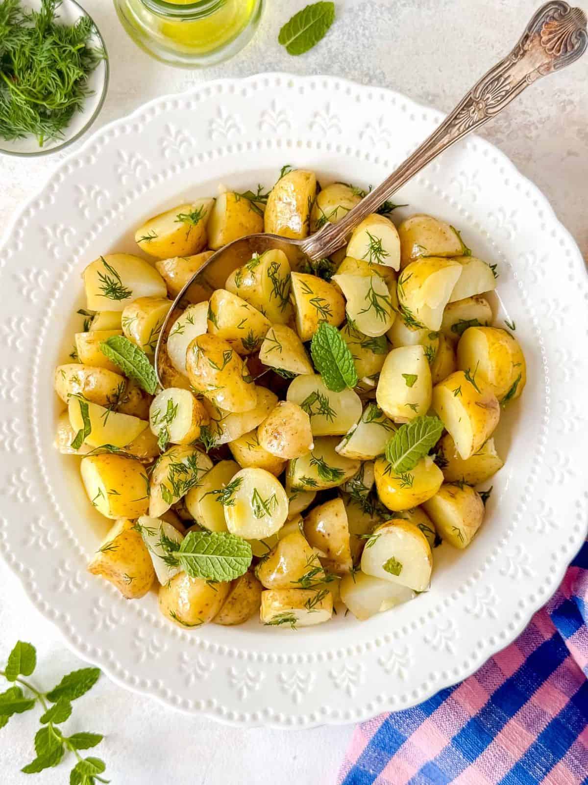 no mayo potato salad with dill and mint in a white bowl next to a bottle of olive oil and a blue and pink cloth.