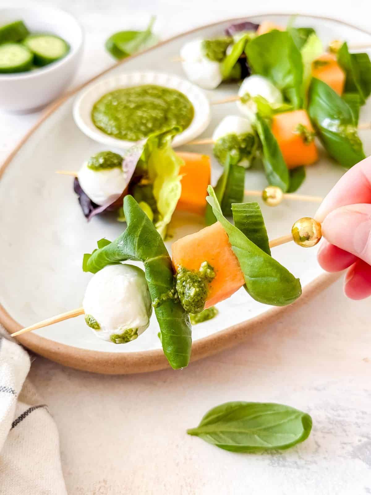 a salad kebab held up with more salad kebabs on a plate in the background, with basil leaves and a small bowl of Persian cucumbers.