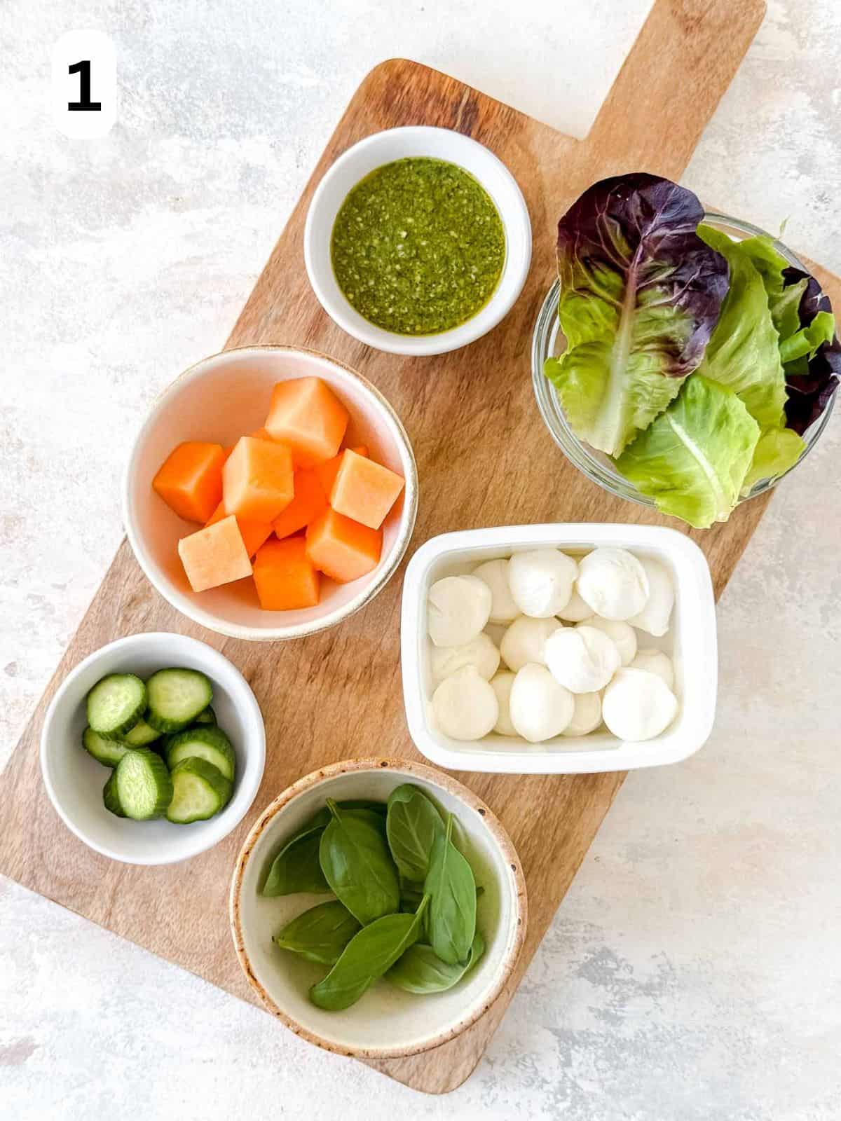 bowls of mini mozzarella balls, lettuce, melon, cucumber, basil leaves and pesto, labelled number one.