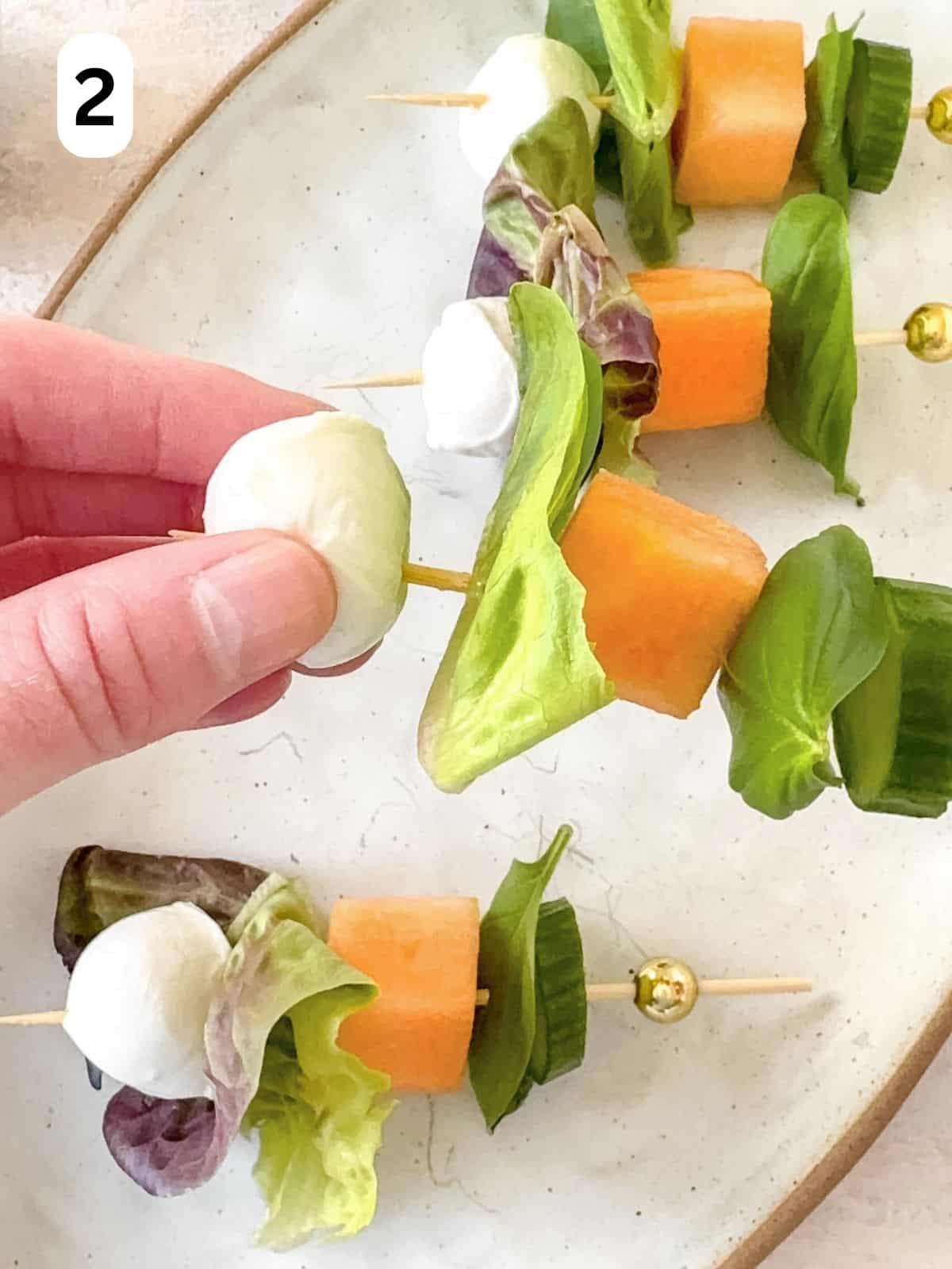 a mini mozzarella ball being threaded onto a kebab with other salad foods, labelled number two.