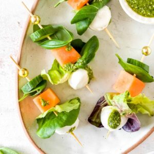 salad kebabs with mozzarella, lettuce, cucumber, melon and basil leaves on a plate with a small bowl of pesto.