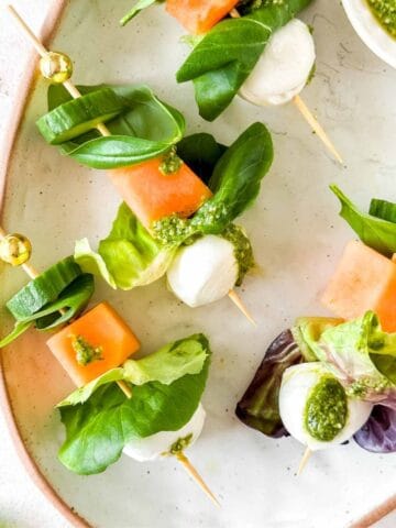 salad kebabs with mozzarella, lettuce, cucumber, melon and basil leaves on a plate with a small bowl of pesto.