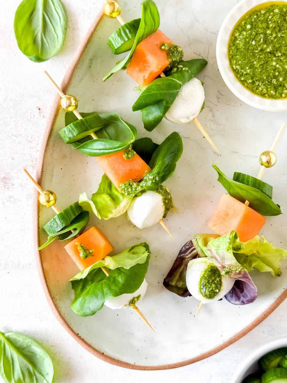 salad kebabs with mozzarella, cucumber, lettuce and melon on a plate next to a small bowl of pesto.