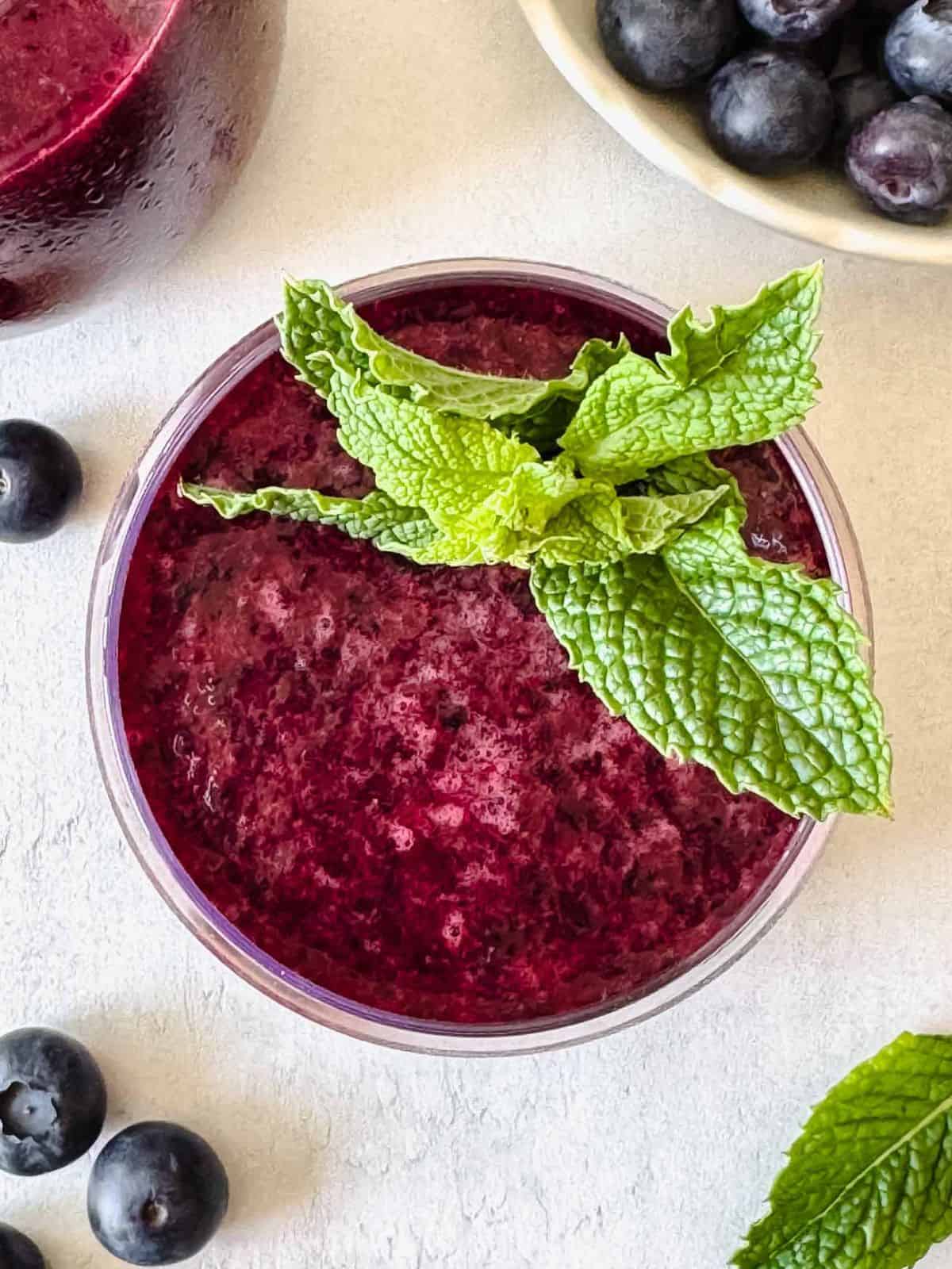 blueberry slushie in a glass topped with fresh mint leaves next to a bowl of blueberries.