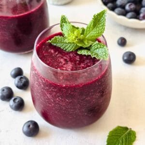 blueberry slushie in a glass garnished with fresh mint leaves next to a bowl of blueberries.