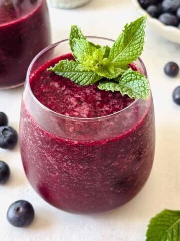 blueberry slushie in a glass garnished with fresh mint leaves next to a bowl of blueberries.
