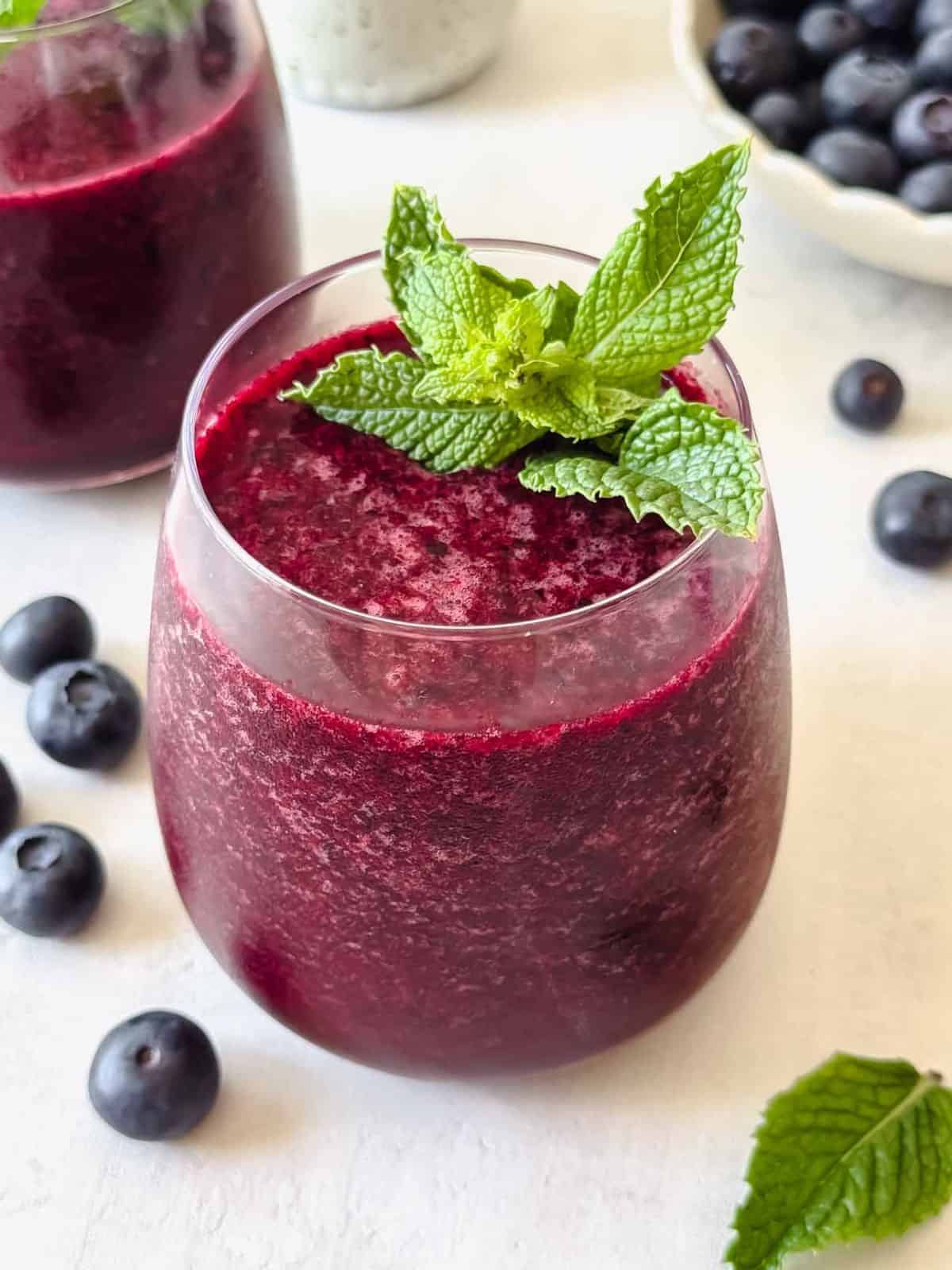 blueberry slushie in a glass next to fresh mint leaves and blueberries.