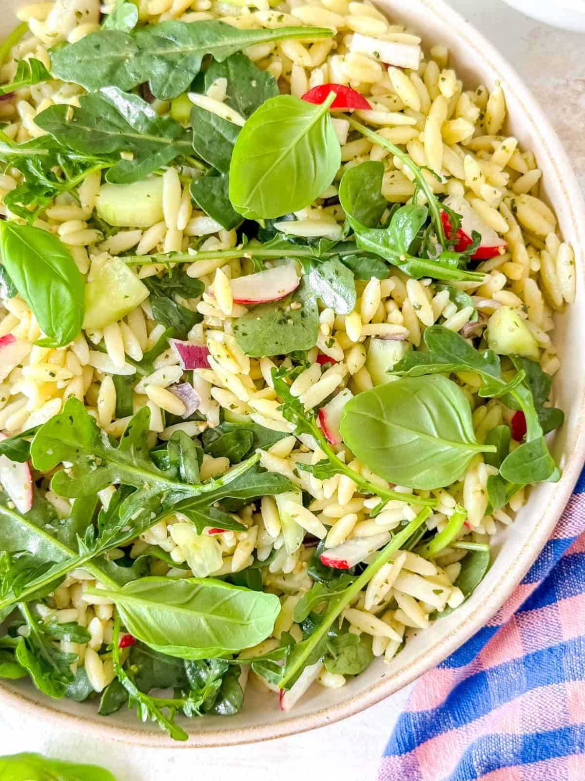 arugula orzo salad in a bowl next to a pink and blue checked cloth.