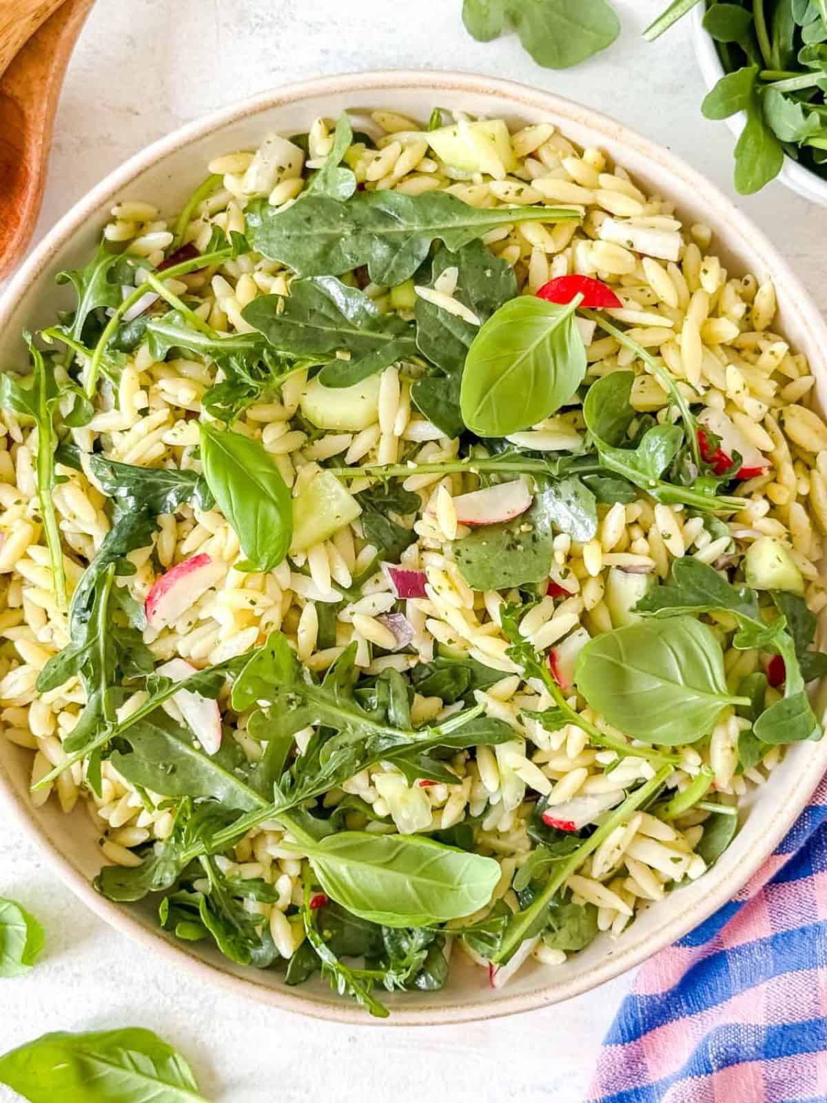 arugula orzo salad in a bowl garnished with fresh basil leaves next to a bowl of arugula leaves and checked cloth.