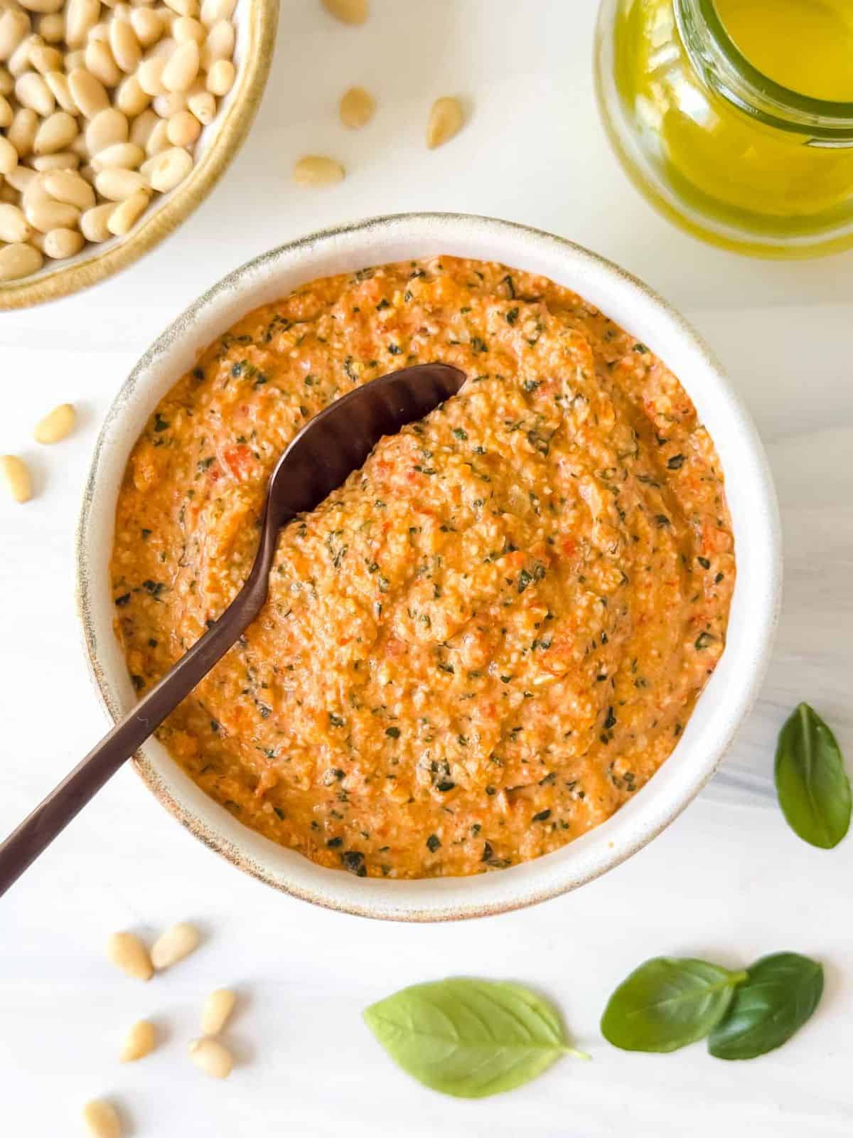 roasted red pepper pesto in a bowl next to fresh basil leaves, olive oil and pine nuts.