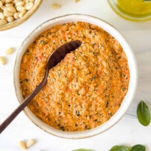 roasted red pepper pesto in a bowl next to pine nuts, fresh basil leaves and olive oil.