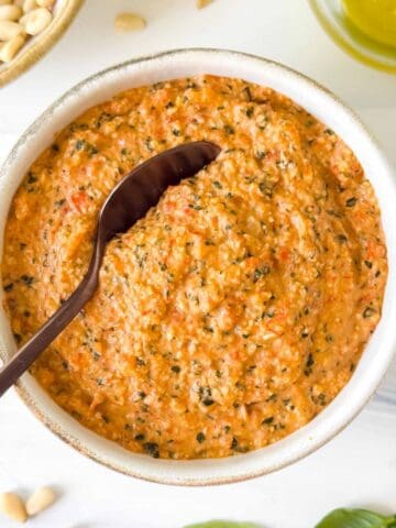 roasted red pepper pesto in a bowl next to pine nuts, fresh basil leaves and olive oil.