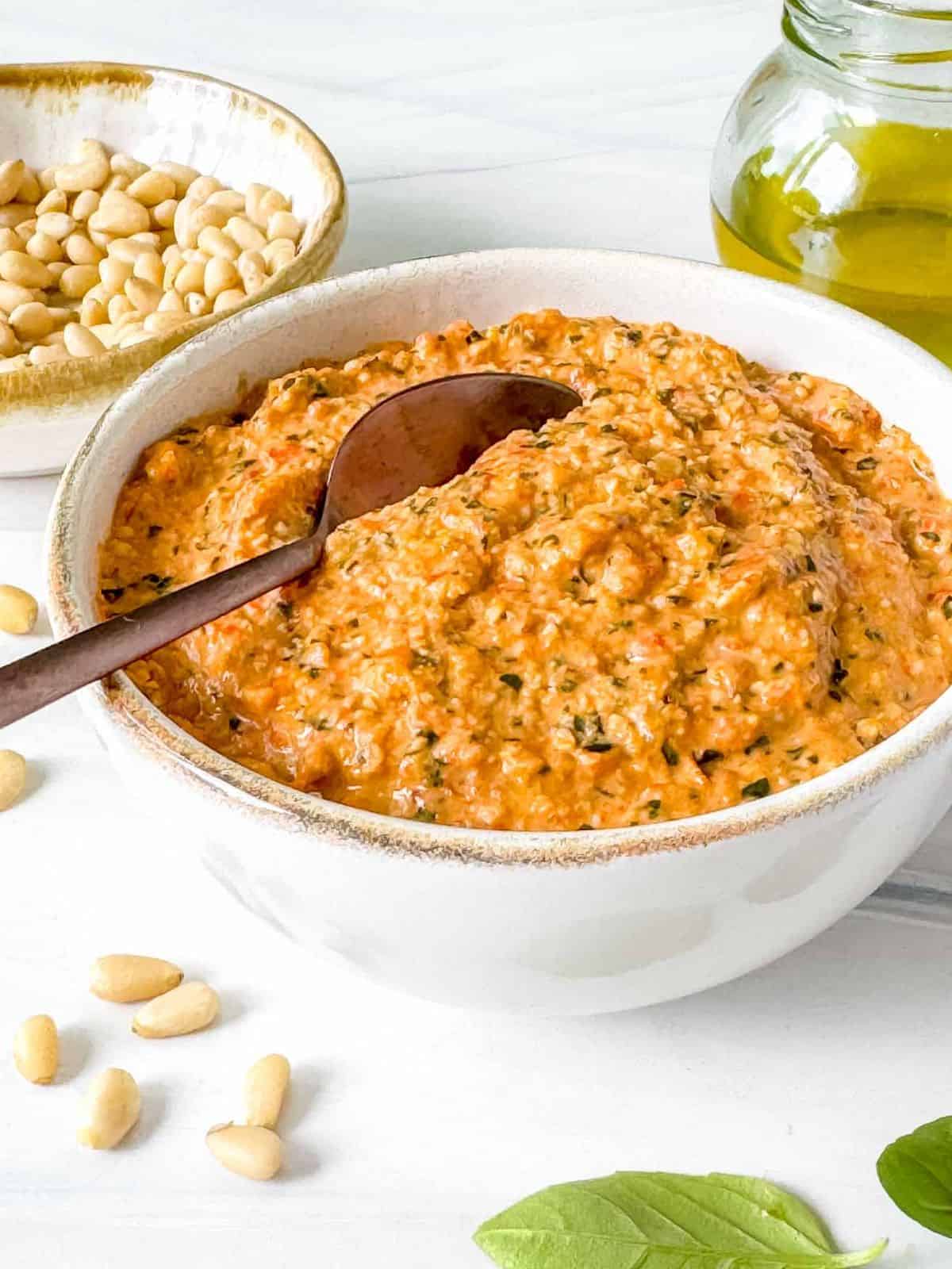 roasted red pepper pesto sauce in a bowl next to olive oil, pine nuts and fresh basil leaves.