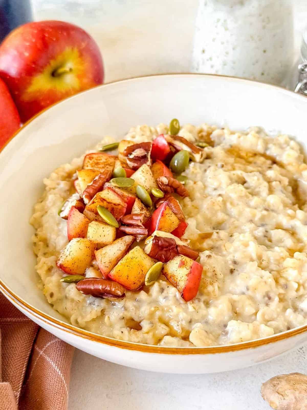 ginger oatmeal in a white bowl next to red apples and topped with pecans, apple and pumpkin seeds.