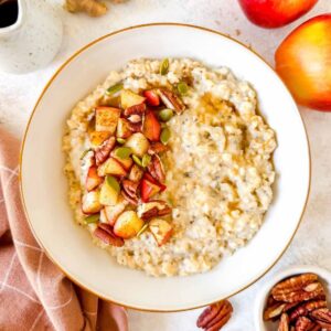 ginger oatmeal in a white bowl topped with pecans, cooked apple and pumpkin seeds.