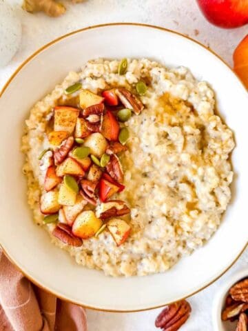 ginger oatmeal in a white bowl topped with pecans, cooked apple and pumpkin seeds.